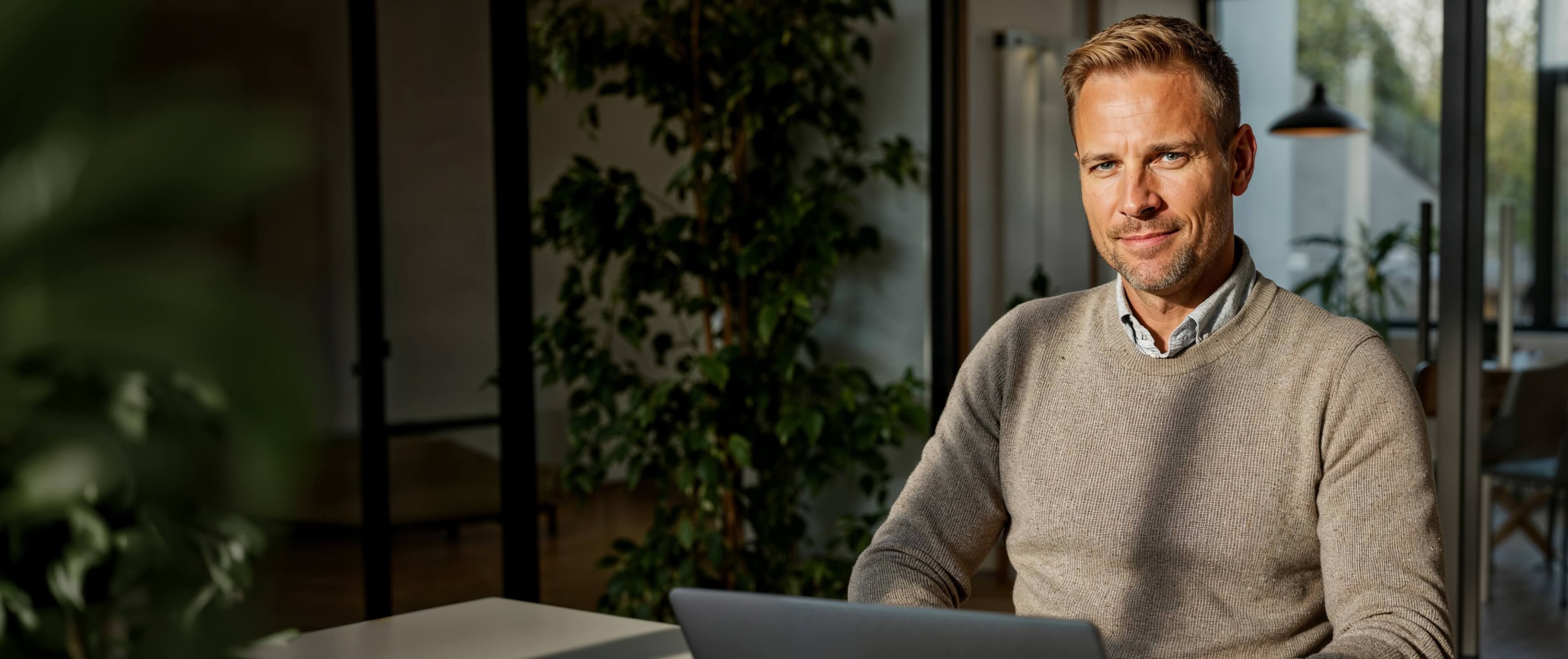A man sits in an office with a laptop, looking directly at the camera.